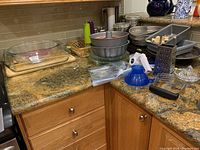 Photo showing assortment of glass baking dishes on countertop, metal baking pans, salt and pepper shakers, kitchen canisters, muffin cups, a blue plastic juicer, metal graters, scissors and cutlery.