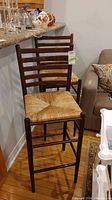 Two wooden bar stools with ladder-back design and woven rush seats positioned next to a counter and sofa, showing overall condition and style.