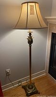 Full view of one brass floor lamp turned on next to a wall, showing fabric shade and floral brass base.