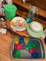 Photo showing green plastic ice bucket, cactus/floral design plates, green & white patterned plates, translucent blue pitcher, and colorful goblets in a gray plastic bin.