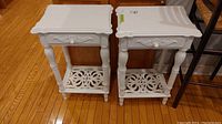 Pair of white painted wood end tables side-by-side on hardwood floor, showing front and side details including drawers and lower shelves.