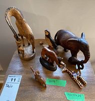 Six wooden wildlife figures displayed on a table: two giraffes on a base, a hippopotamus, elephant, puffin with beak detached, a smaller fox, and one other small animal figurine.