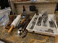 Overview of various kitchen utensils spread on countertop showing rolling pin, wooden spoons, metal tongs, and white cutlery tray filled with stainless steel flatware