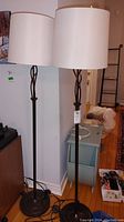 Two metal floor lamps with white drum shades standing side by side against a wall, hardwood floor setting.
