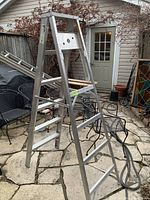 Full view of metal 6-foot step ladder standing outdoors on patio stone floor next to garden furniture.