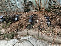 Seven black outdoor solar spotlights placed in garden soil with surrounding dried leaves and plants, showing solar panels and spotlight heads