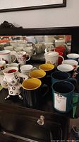 Photo showing multiple assorted mugs including dark blue with yellow interior, white with prints, and novelty shaped mug on a black piece of furniture