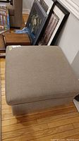 Beige upholstered square ottoman sitting on wood floor with framed art leaning against wall in background.
