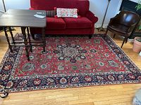 Full view of the floral motif area rug with a red central area and a navy and cream floral border, placed on wooden floor under a red couch and wooden tables.