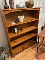 Front view of pine shelving unit with items on shelves removed except for photo context. Four fixed shelves visible. Wood grain and finish tone visible. Light wear and scratches noted.