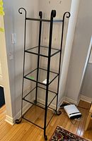 Front view of five tiered shelving unit with clear glass shelves and black metal frame with curled top corners and curled feet.