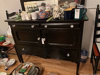 Front view of the black wooden sideboard with the mirrored backsplash, three drawers on the top, and one cabinet door in the middle. The sideboard is adorned with various mugs and glass knobs on the drawer handles.