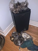 Collection of silver plated tableware including a gravy boat, sugar bowl, creamer, pepper shakers, placed on a speaker with additional glassware around it.
