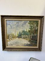 Front view of the framed oil painting showing street scene with figures, greenery, wall, and cloudy sky. Visible artist signature on bottom left.