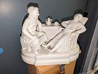 White bisque porcelain figural sculpture with a laughing priest reclining in chair and another man seated on stool, tea set on table behind.