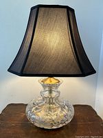 Front view of the illuminated lamp showing the black fabric shade and cut crystal base on wood surface.