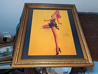 Framed artwork featuring a pin-up style illustration of a woman with red hair, black high heels, and a black corset against a yellow background.