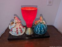 Front view of the mid-century modern table lamp with two figural ladies and red cone fiberglass lampshade on a black base against neutral wall