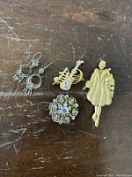 Four jewelry pieces including 925 silver earrings, rhinestone guitar and floral brooches, and a gold-tone Art Deco lady brooch on a wooden surface.