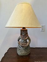 Front view of the Mid-Century Modern West German figural lady art pottery table lamp with fabric shade on a wooden table against a plain wall.