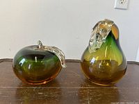 Pair of Murano glass pear and apple bookends in green and amber gradient, with silver-colored glass leaves. Apple missing top stem. Images show glossy surface and size relative to hand.
