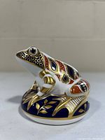 Side view showing the detailed gold, red, and blue pattern on the frog sitting on a cushion.