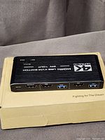 Photo of the black rectangular 4K Ultra HD HDMI USB KVM switch with three HDMI input ports and multiple USB ports placed on tan box.