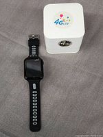 Black W5 smart watch with rectangular touchscreen and black strap with white circular ventilation holes, placed beside its white 4G LTE branded box.