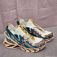 Side profile showing the intricate beige blade-style sole, knit upper in cream with teal overlays, and patterned orange accents.