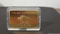 Front view showing inscription '100 Mills .999 Fine Gold 1 Troy Ounce' and a raised bear image on the gold plated bar in clear plastic case.