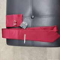 Photo showing a burgundy tie, matching pocket square, silver tie clip and pair of cufflinks laid out on black surface.