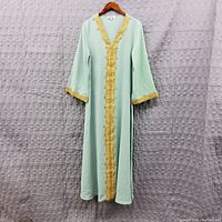 Photo of a light green women's kaftan with gold fringe trim along front opening and sleeve edges, hanging on a wooden hanger against a grey textured background.