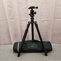 K&F Concept tripod set up upright with black carrying case placed horizontally behind it, showing tripod legs, foam grip, and ball head mount with adjustment knobs.