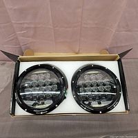 Pair of round LED headlights inside cardboard box with protective foam insert.