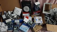 Box with assorted new household electrical items including multiple circuit breaker boxes, switches, sockets, lighting components, and wiring accessories visible.