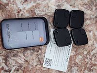Photo showing four black square Smart Finder location trackers and one black rectangular remote control device with instruction cards on a surface.