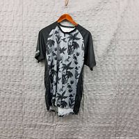 Front view of a men's size L t-shirt showing black sleeves and gray body with tropical-themed black print including palm trees, flamingos, and bicycles.