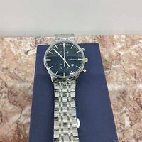 Wanvi watch face close-up showing black dial with silver hands, chronograph subdials, date window, and silver metal bracelet.