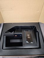 Black wireless cellphone car charger sitting in a cardboard box, showing charging pad with wireless symbol and a small digital or LED display.