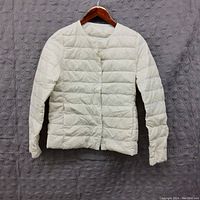 Front view of a white women's puffer jacket hanging against a textured wall, showing full length and details like snap buttons and pockets.