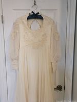 Full front view of the ivory 1970s wedding dress hanging on a door, showing lace decorations around the neckline and shoulders and sheer long sleeves with lace cuffs.