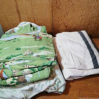 Folded green flannel double bed sheet set with knight on horse, castle and dragon print next to folded white duvet cover with grey stripes.