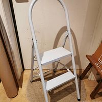 White two-step folding metal ladder standing on floor, showing overall structure and condition.