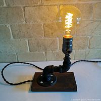 Full view of the industrial style pipe lamp with Edison filament bulb lit on a solid wooden base and plugged in.