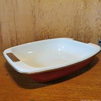 Large KitchenAid lasagna or baking pan with red exterior, white ceramic interior, rectangular shape, and dual handles, displayed on a wooden surface.