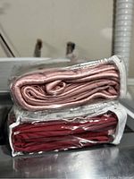 Two bed sheets folded and sealed in clear plastic bags: one light pink and one red, displayed on a metal surface.