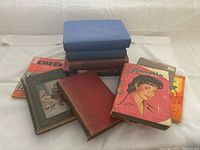 Overhead image showing a stack of nine antique and vintage hardcover books in various colors including red, blue, green, and orange with visible wear and vintage illustrations on some covers.