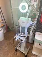 Full machine showing Catena branded steamer on wheeled stand with attached magnifying lamp, cords and handheld components visible, machine positioned in a room with tile floor.