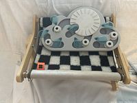 Photo of a gray cat treat puzzle toy with blue rotating components resting on a wooden framed cat window swing with black and white checkered fabric.