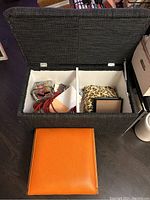 Open storage bench showing internal divided compartments holding various items like cords and fabric pieces. Orange footstool positioned in front.